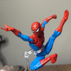 May include: A Spider-Man action figure in a dynamic pose, featuring a red and blue costume. The figure has a detailed web pattern on the red sections, with a white-eyed mask. The figure is supported by a clear stand, showcasing its acrobatic stance.