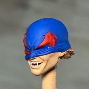 May include: A painted miniature head sculpt featuring a blue mask with red eye markings. The face has a light skin tone, a wide grin revealing white teeth with pointed fangs, and visible brown hair at the back. The head is mounted on a beige post.