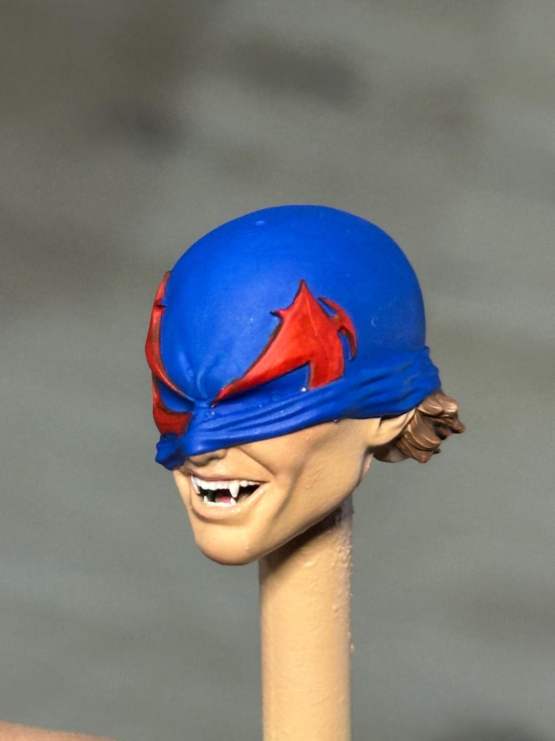 May include: A painted miniature head sculpt featuring a blue mask with red eye markings. The face has a light skin tone, a wide grin revealing white teeth with pointed fangs, and visible brown hair at the back. The head is mounted on a beige post.