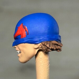 May include: A miniature figurine head featuring a bright blue helmet with a red emblem on the side. The face is painted with a light skin tone, showing bared teeth and a snarling expression. Brown hair is visible at the base of the blue helmet.