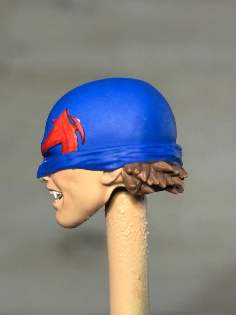 May include: A miniature figurine head featuring a bright blue helmet with a red emblem on the side. The face is painted with a light skin tone, showing bared teeth and a snarling expression. Brown hair is visible at the base of the blue helmet.