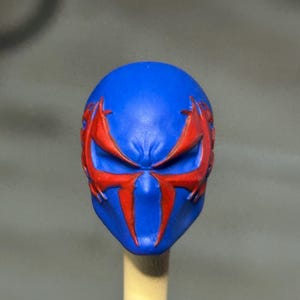 May include: A blue sculpted head piece featuring a prominent red spider-like emblem that extends across the eye area. The design includes sharp, angular lines and a smooth matte finish, with the mask mounted on a light-colored cylindrical support.