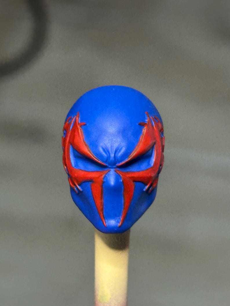 May include: A blue sculpted head piece featuring a prominent red spider-like emblem that extends across the eye area. The design includes sharp, angular lines and a smooth matte finish, with the mask mounted on a light-colored cylindrical support.