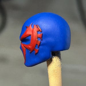 May include: A blue sculpted head piece featuring a prominent red stylized spider-like emblem on the side. The mask has a smooth matte finish and a distinct, sharp-edged design around the eye area, mounted on a vertical support post.