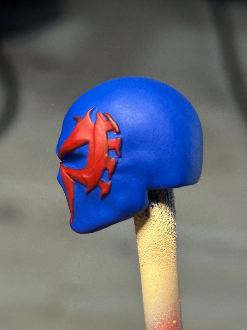 May include: A blue sculpted head piece featuring a prominent red stylized spider-like emblem on the side. The mask has a smooth matte finish and a distinct, sharp-edged design around the eye area, mounted on a vertical support post.