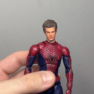 Może przedstawiać: Figurka Spider-Mana w klasycznym czerwono-niebieskim kostiumie. Figurka ma szczeg&oacute;łowy wz&oacute;r pajęczyny na czerwonych częściach i czarny emblemat pająka na klatce piersiowej. Figurka jest trzymana w dłoni.