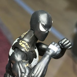 May include: A black action figure with a white spider emblem on the chest and white eye patches. The figure is in a fighting stance with clenched fists, showing visible wear and paint damage on the shoulder and upper arm joints.