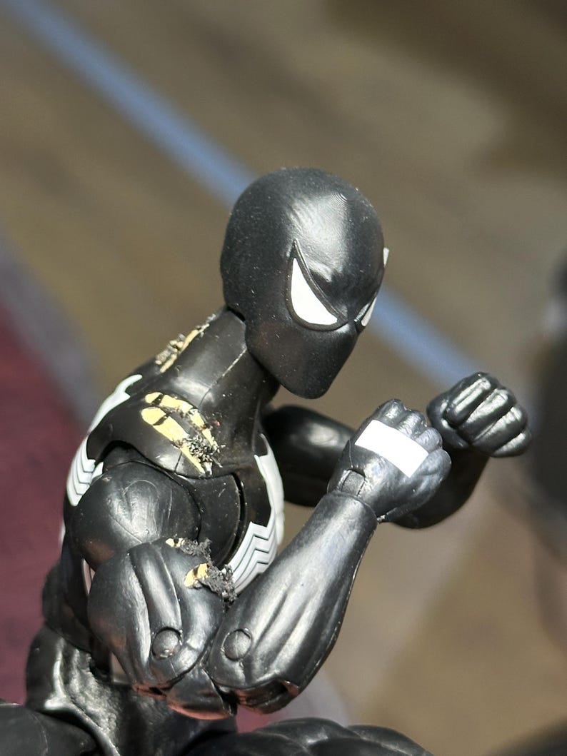 May include: A black action figure with a white spider emblem on the chest and white eye patches. The figure is in a fighting stance with clenched fists, showing visible wear and paint damage on the shoulder and upper arm joints.