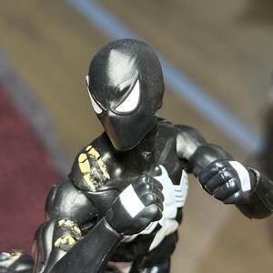 May include: A black action figure with a white spider emblem on the chest and white eye patches. The figure features articulated joints, white rectangular markings on the wrists, and visible paint wear on the shoulder and arm areas.