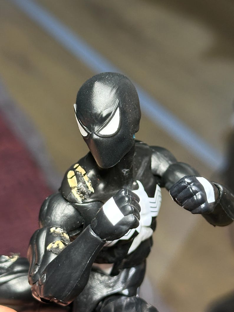 May include: A black action figure with a white spider emblem on the chest and white eye patches. The figure features articulated joints, white rectangular markings on the wrists, and visible paint wear on the shoulder and arm areas.