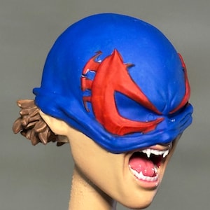 May include: A sculpted figurine head featuring a bright blue mask with prominent red eye-shaped accents. The face has an open mouth revealing white fangs and a pink tongue. Brown hair is visible at the back, and the neck is a plain, unpainted beige cylinder.