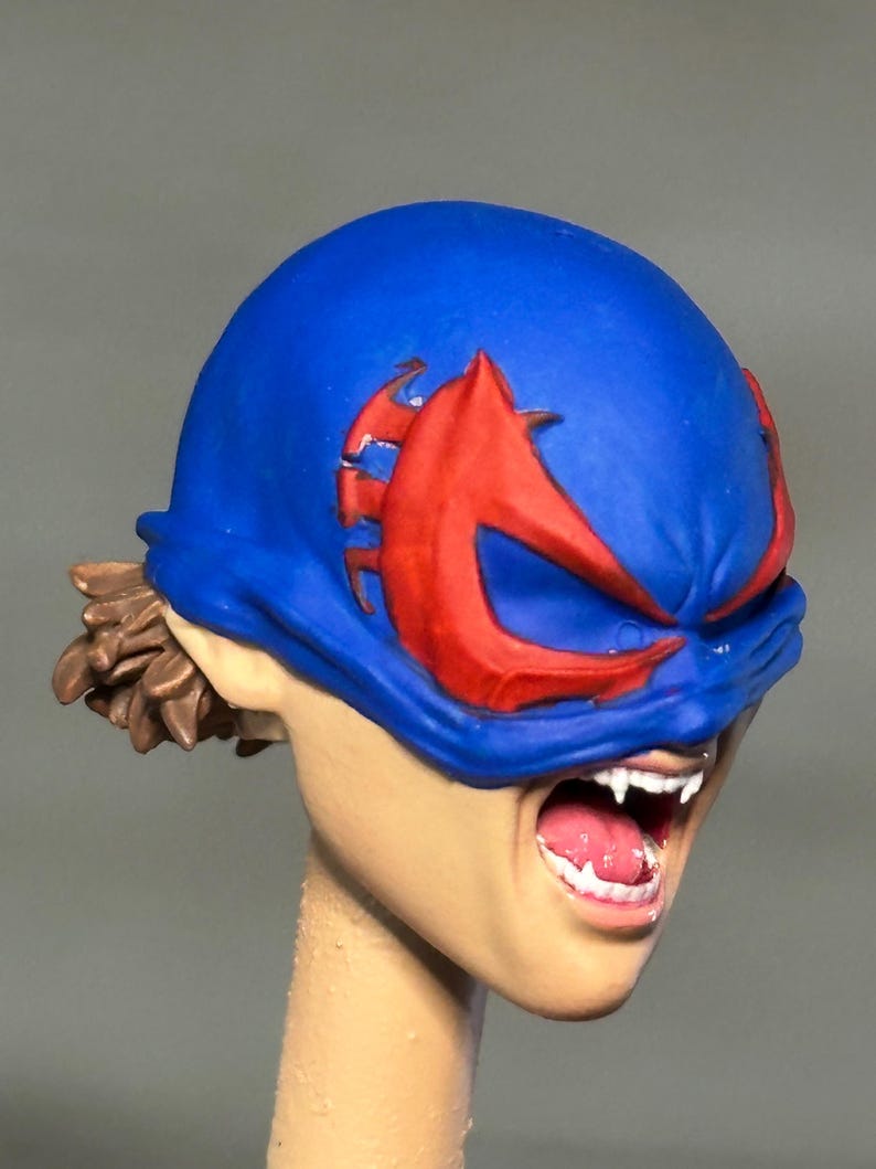 May include: A sculpted figurine head featuring a bright blue mask with prominent red eye-shaped accents. The face has an open mouth revealing white fangs and a pink tongue. Brown hair is visible at the back, and the neck is a plain, unpainted beige cylinder.
