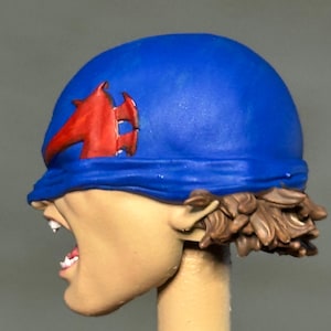 May include: A painted miniature head featuring a blue head covering with a red emblem on the side. The figure has light skin, an open mouth showing white teeth, and brown hair visible at the base of the blue head covering, mounted on a post.