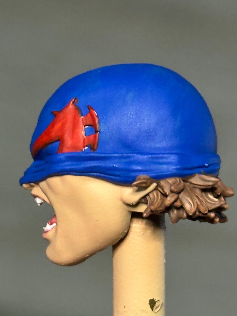 May include: A painted miniature head featuring a blue head covering with a red emblem on the side. The figure has light skin, an open mouth showing white teeth, and brown hair visible at the base of the blue head covering, mounted on a post.