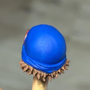 May include: A miniature figurine head featuring a bright blue bandana wrapped around the crown. The hair is sculpted in short, spiky brown layers that peek out from beneath the fabric. A small, circular red emblem is visible on the side of the blue bandana.