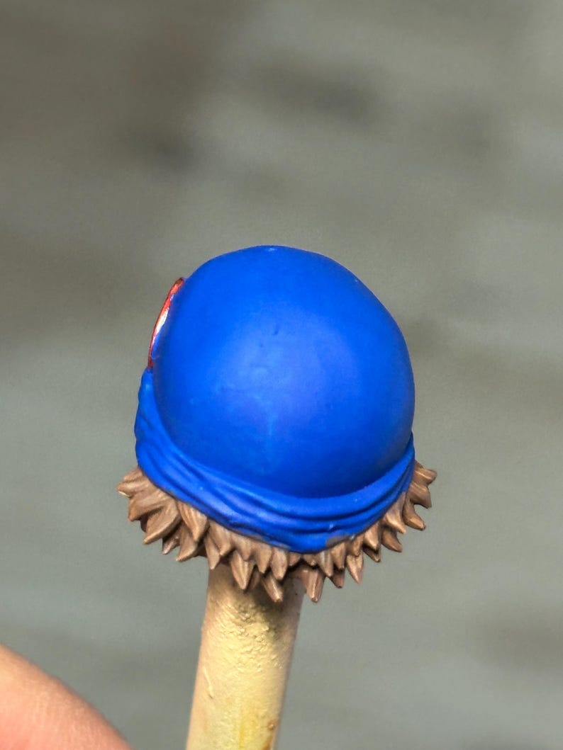 May include: A miniature figurine head featuring a bright blue bandana wrapped around the crown. The hair is sculpted in short, spiky brown layers that peek out from beneath the fabric. A small, circular red emblem is visible on the side of the blue bandana.