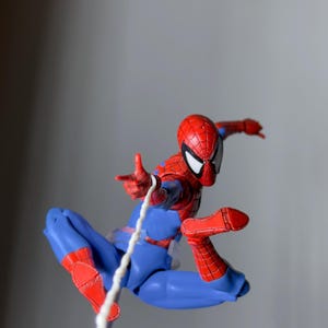 Puede incluir: Figura de acci&oacute;n de Spider-Man en pose din&aacute;mica, con un traje rojo y azul con detalles de telara&ntilde;a. La figura est&aacute; en el aire, con una telara&ntilde;a blanca que se extiende desde su mano. La m&aacute;scara tiene lentes blancas.