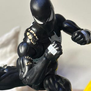 May include: A black action figure with white eye lenses and a white chest emblem. The figure features articulated joints, white accents on the hands, and visible wear with chipped paint revealing a light-colored base layer on the shoulder and arm.
