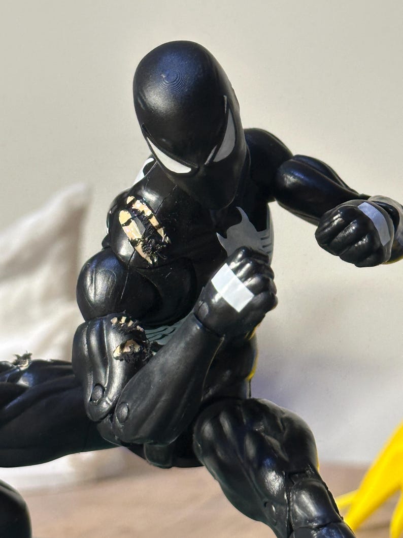 May include: A black action figure with white eye lenses and a white chest emblem. The figure features articulated joints, white accents on the hands, and visible wear with chipped paint revealing a light-colored base layer on the shoulder and arm.