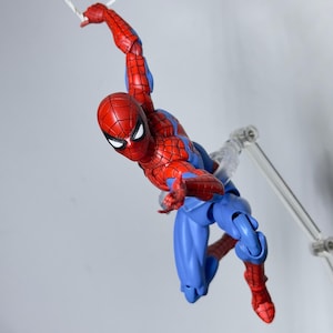 Puede incluir: Figura de acci&oacute;n de Spider-Man en rojo y azul en una pose din&aacute;mica, balance&aacute;ndose de una telara&ntilde;a blanca. La figura tiene un disfraz detallado y est&aacute; sostenida por un soporte transparente, sobre un fondo liso.
