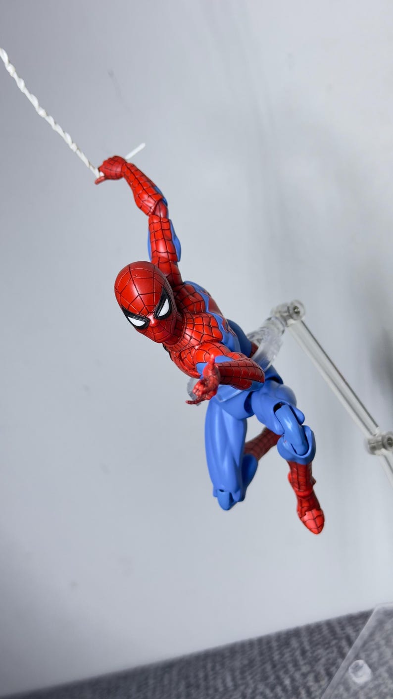 Puede incluir: Figura de acci&oacute;n de Spider-Man en rojo y azul en una pose din&aacute;mica, balance&aacute;ndose de una telara&ntilde;a blanca. La figura tiene un disfraz detallado y est&aacute; sostenida por un soporte transparente, sobre un fondo liso.