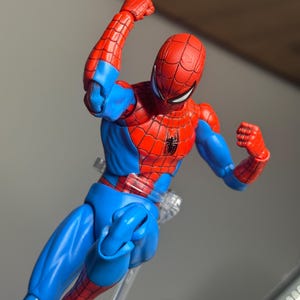 May include: A red and blue Spider-Man action figure in a dynamic pose. The figure features a red mask and suit with blue accents, and a black spider emblem on the chest. The figure is supported by a clear stand.