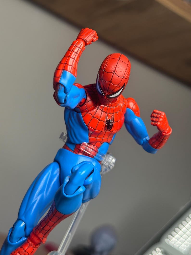 May include: A red and blue Spider-Man action figure in a dynamic pose. The figure features a red mask and suit with blue accents, and a black spider emblem on the chest. The figure is supported by a clear stand.