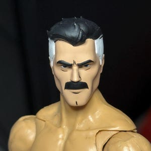 Può includere: Una testa di action figure in plastica raffigurante un personaggio dalla pelle abbronzata, con folti baffi neri e capelli neri con i lati bianchi. Il personaggio presenta lineamenti definiti, tra cui sopracciglia marcate e una piccola linea nera sul mento.