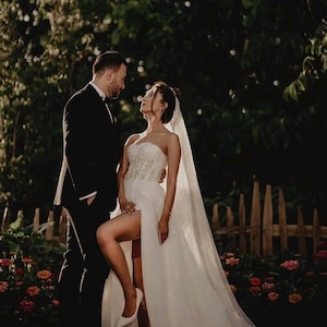 May include: A couple poses for a wedding photograph. The bride wears a white strapless gown with a high leg split and a veil. The groom is in a black tuxedo. The background features a wooden fence, flowers, and foliage. The photo is taken at dusk.