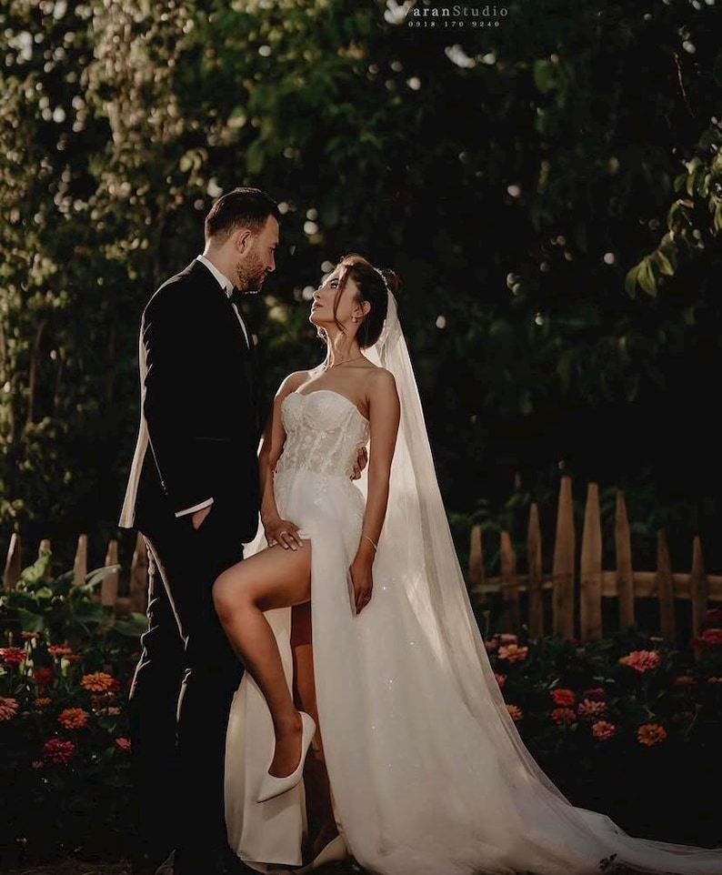 May include: A couple poses for a wedding photograph. The bride wears a white strapless gown with a high leg split and a veil. The groom is in a black tuxedo. The background features a wooden fence, flowers, and foliage. The photo is taken at dusk.