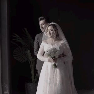 May include: A bride in a white lace wedding dress with long sleeves and a veil, holding a bouquet of white flowers. A man in a dark suit stands behind her, embracing her. The setting is a dimly lit room with a large plant.