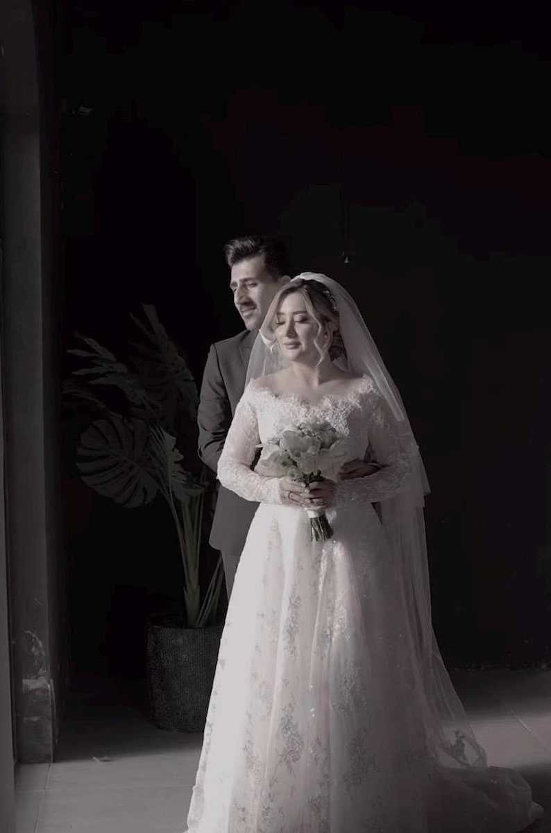 May include: A bride in a white lace wedding dress with long sleeves and a veil, holding a bouquet of white flowers. A man in a dark suit stands behind her, embracing her. The setting is a dimly lit room with a large plant.