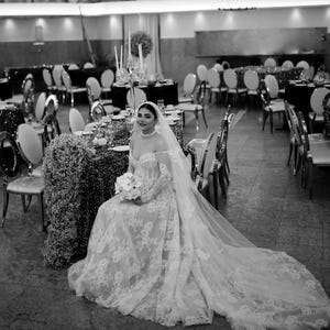 May include: A bride in a white lace wedding dress with a long train and veil sits at a decorated table. She holds a bouquet of white flowers. The setting is a formal event with tables and chairs.
