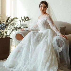 May include: A white wedding dress with an off-the-shoulder neckline and long, lace sleeves. The bride is wearing a veil and seated on a grey armchair. The dress has a full skirt and is adorned with delicate lace, creating a romantic look.