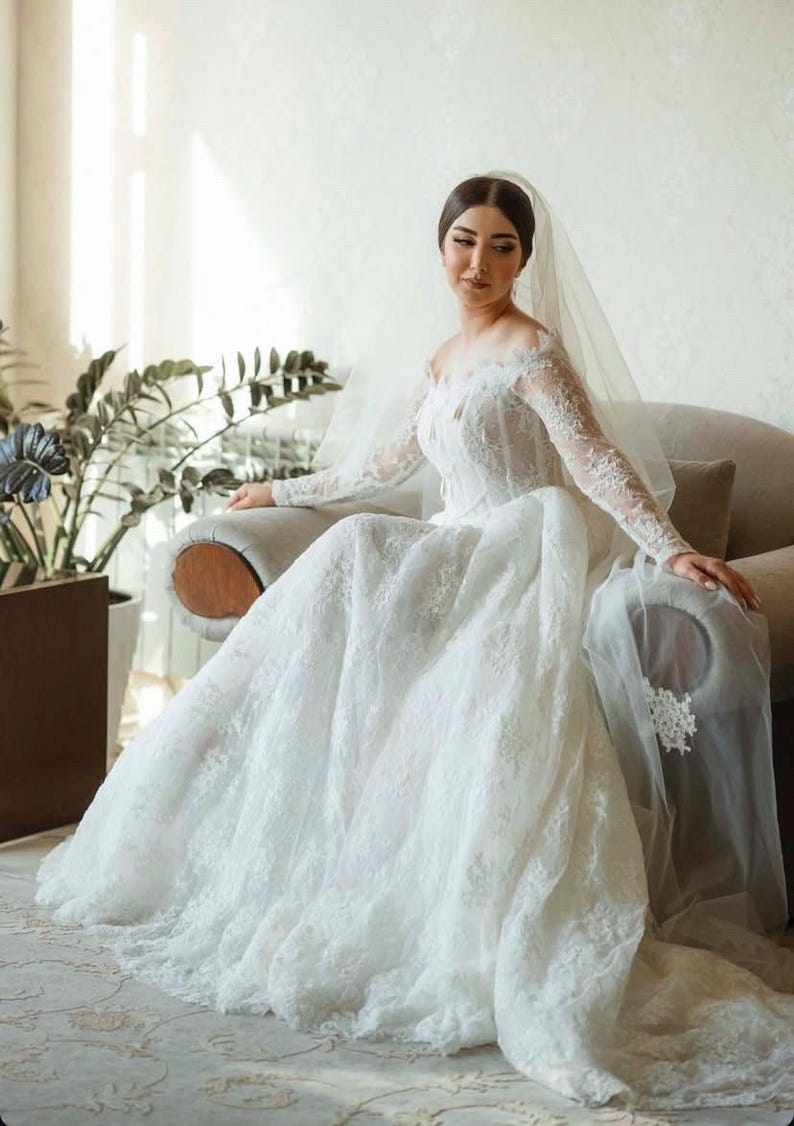 May include: A white wedding dress with an off-the-shoulder neckline and long, lace sleeves. The bride is wearing a veil and seated on a grey armchair. The dress has a full skirt and is adorned with delicate lace, creating a romantic look.