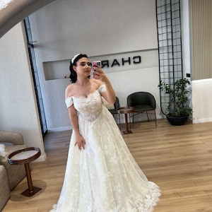 May include: A white off-the-shoulder wedding dress with a sweetheart neckline and a full lace skirt. The dress features intricate lace detailing and a flowing silhouette. The dress is captured in a mirror.
