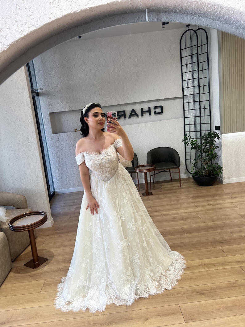 May include: A white off-the-shoulder wedding dress with a sweetheart neckline and a full lace skirt. The dress features intricate lace detailing and a flowing silhouette. The dress is captured in a mirror.