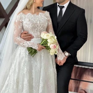 May include: A bride in a white lace wedding dress with long sleeves and a veil, holding a bouquet of white and pink flowers. A groom in a black suit and tie stands beside her, both smiling. The wedding dress features an off-the-shoulder neckline.