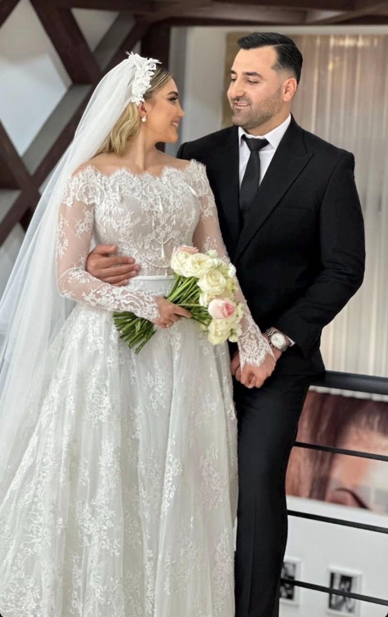 May include: A bride in a white lace wedding dress with long sleeves and a veil, holding a bouquet of white and pink flowers. A groom in a black suit and tie stands beside her, both smiling. The wedding dress features an off-the-shoulder neckline.
