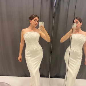 May include: A white, strapless, floor-length gown with detailed beading. The dress has a fitted shape and a small slit. The image shows the dress in a fitting room, with a mirror reflecting the dress.