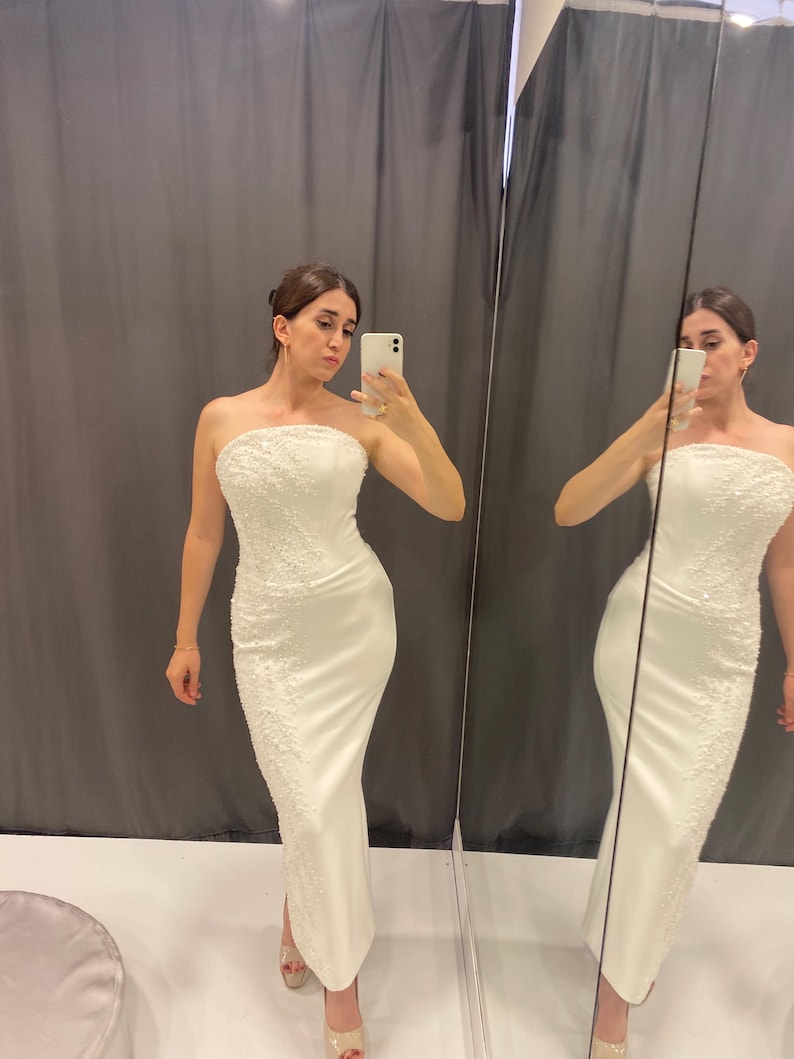 May include: A white, strapless, floor-length gown with detailed beading. The dress has a fitted shape and a small slit. The image shows the dress in a fitting room, with a mirror reflecting the dress.