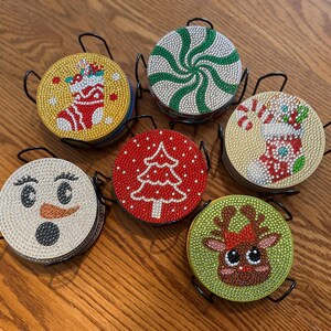 May include: Set of six Christmas-themed coasters with black metal holders. Each coaster features a different design: a snowman, a Christmas tree, a reindeer, a stocking, and a peppermint swirl. The coasters are embellished with small, sparkling gems.