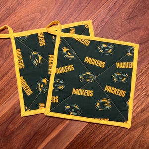 May include: Two green and yellow Packers-themed pot holders. The pot holders have a dark green background with a repeating pattern of the Packers logo and the word "PACKERS" in yellow. They have yellow trim and a loop for hanging.