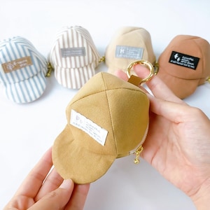 Puede incluir: Una colección de pequeños monederos con forma de gorra en varios colores, incluyendo rayas azules y blancas, beige y marrón. Se sostiene un monedero, mostrando una cremallera dorada y una pequeña etiqueta con texto. Los monederos tienen un diseño de gorra de béisbol.