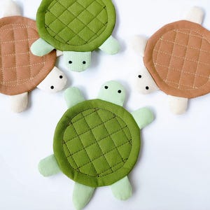 May include: Four turtle-shaped coasters. The coasters have quilted, circular tops in shades of green and brown, with matching turtle feet. The turtle heads are off-white with black dot eyes. These decorative coasters are perfect for a beach or ocean-themed home.