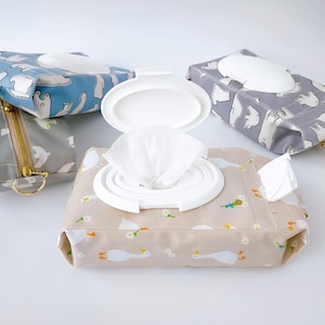 May include: Several reusable baby wipe cases in various colors and patterns. One case is open, revealing white wipes. The cases feature designs with bears, geese, and floral patterns. The cases have a white plastic opening for easy access to the wipes.