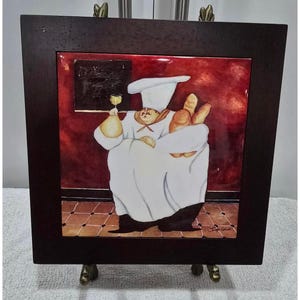 May include: A square decorative tile framed in dark wood featuring a painted chef in a white uniform and tall hat. The chef holds a glass of white wine and a bundle of bread loaves against a deep red background with a tiled floor and black sign.