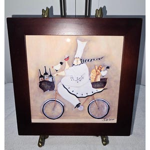 May include: A square ceramic tile framed in dark brown wood featuring a whimsical illustration of a chef in a white uniform riding a bicycle. The chef carries baskets filled with wine bottles, bread, and a small bird, signed by Jennifer Garant.
