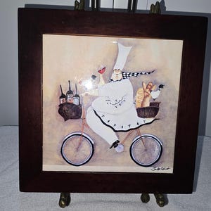 May include: A square ceramic tile framed in dark wood featuring a painting of a chef in a white uniform and tall hat riding a bicycle. The chef carries baskets filled with wine bottles, bread, and a small bird. The artist signature is visible.