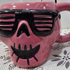 May include: Pink ceramic mug shaped like a skull wearing black sunglasses. The mug has a large handle and a smiling mouth design. A unique and quirky addition to any kitchen or coffee collection.